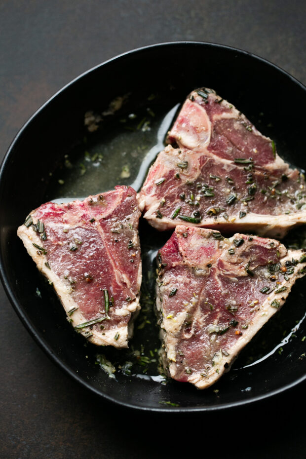 Raw lamb loin chops covered in a marinade in a black bowl.