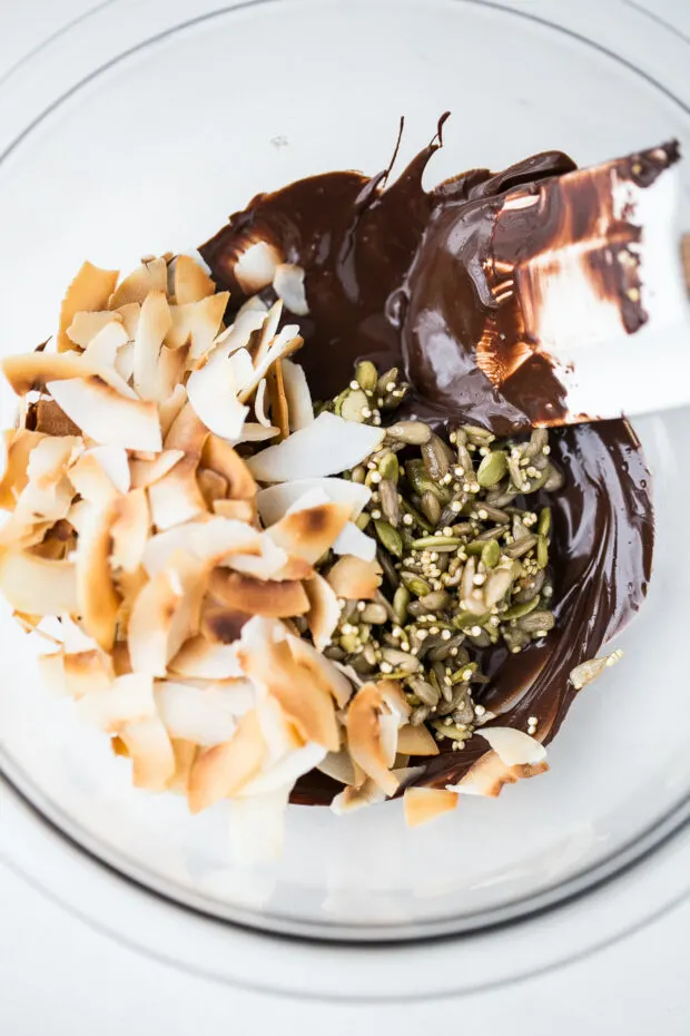 A mixing bowl with melted chocolate on the bottom. Toasted coconut and sticky toasted nuts are on top of the chocolate.