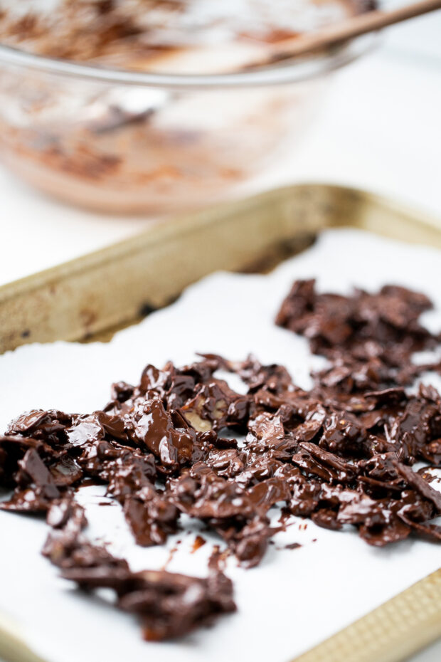 Toasted nuts and seeds covered in melted chocolate are spread on a parchment paper lined pan.
