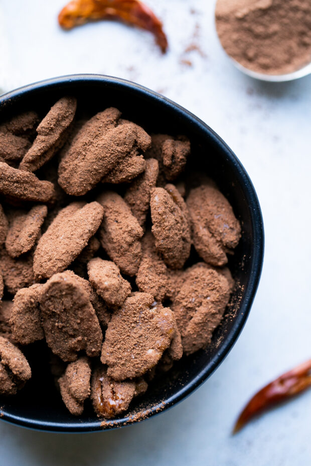 Finished candied pecans with cocoa chile powder in a small black bowl. A spoonful of cocoa powder and a dried chile pepper is in the background.