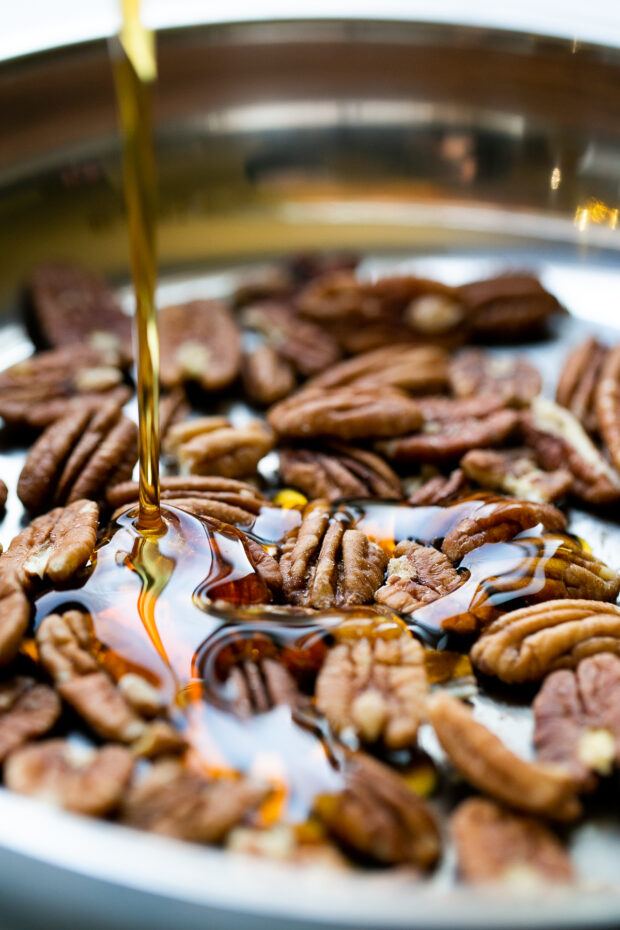 Raw pecans in a skillet with maple syrup being poured over them.
