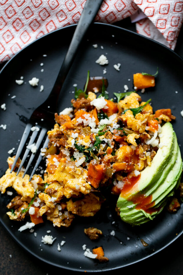 Black plate with sweet potato hash with chorizo and sliced avocado on the side. Hash is topped with a sprinkle of cotija cheese and hot sauce.