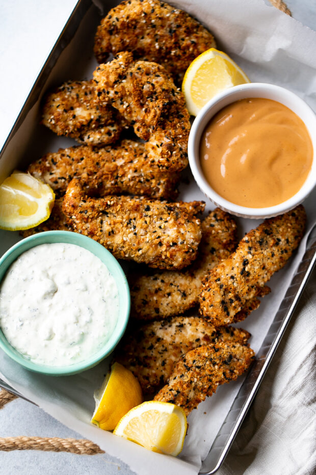 Everything Bagel Chicken Tenders fresh from the air-fryer in a rectangle tin with some dipping sauces and lemon wedges.