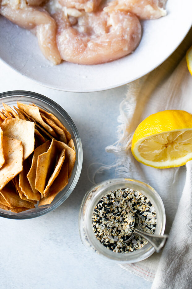 A bowl of raw chicken tenders that have been salted next to a squeezed lemon half, a bowl of crackers, and a container of everything bagel seasoning.