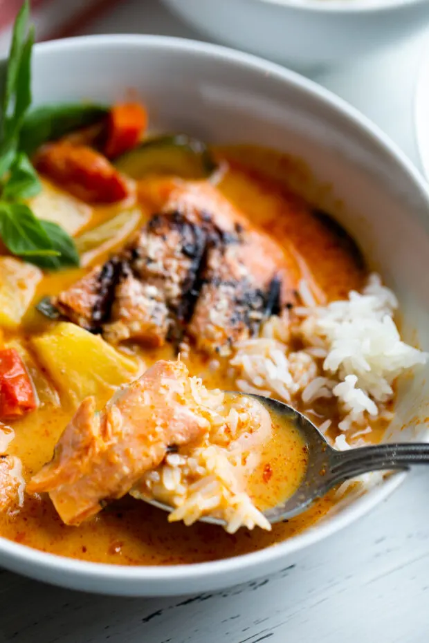A serving bowl with pineapple salmon curry served over jasmine rice. A spoon is grabbing a bite out of the bowl.