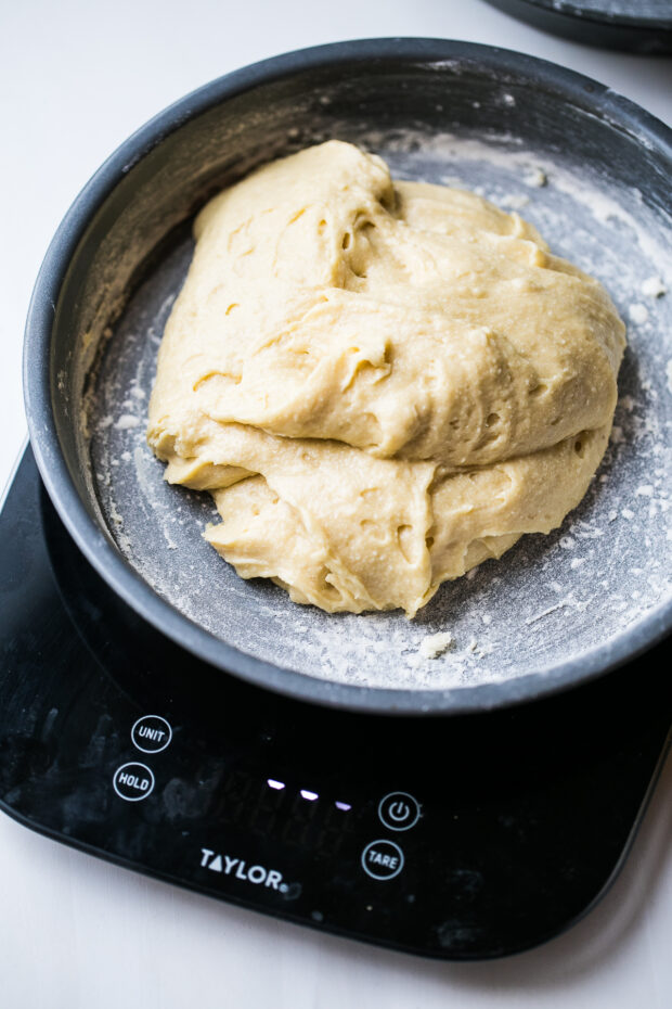 Thick vanilla cake batter in a buttered and floured round cake pan. Pan is sitting on a kitchen scale.