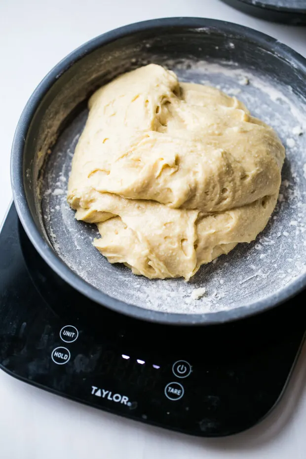 Thick vanilla cake batter in a buttered and floured round cake pan. Pan is sitting on a kitchen scale.