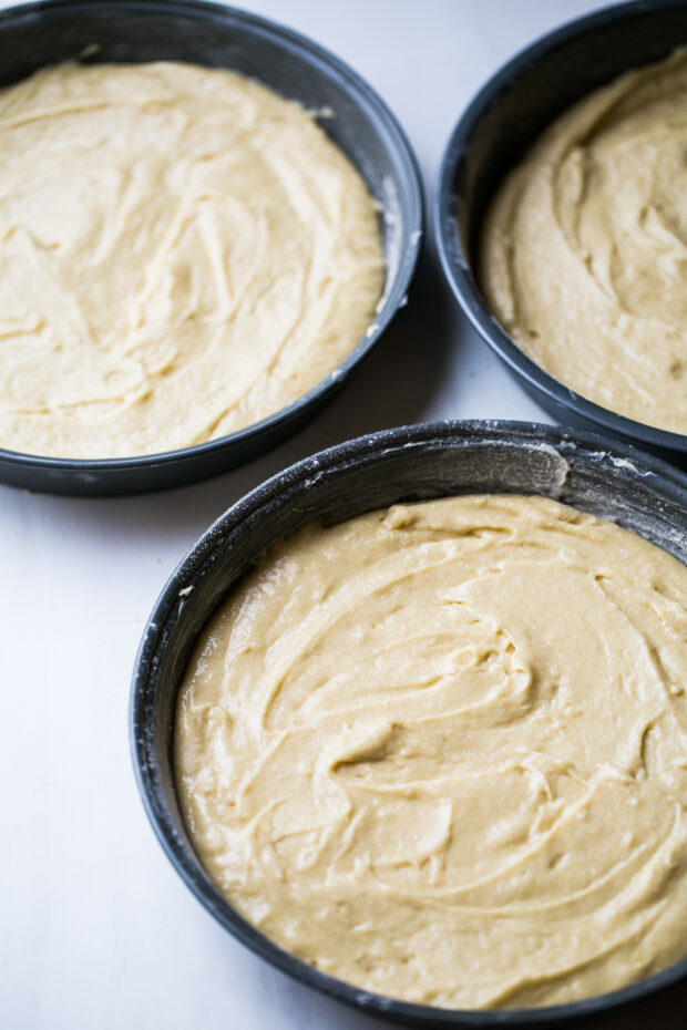 Three buttered and floured cake pans with cake batter spread evenly in them.