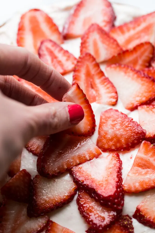 A hand arranging fresh strawberry slices on a frosted cake layer.