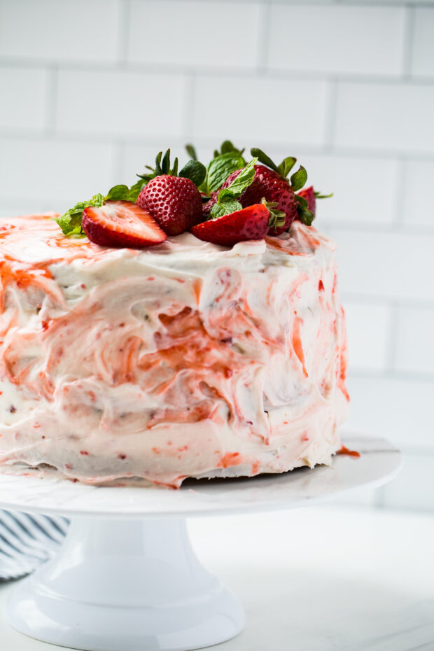 Finished vanilla layer cake with strawberry filling on a white marble cake platter topped with fresh strawberries and mint leaves. 