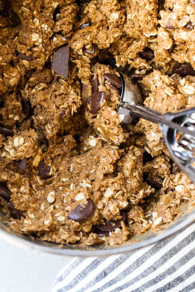 Mixing bowl with Gluten-Free Peanut Butter Oatmeal Cookie dough with chunks of chocolate melts. A cookie scoop is stuck inside the dough.