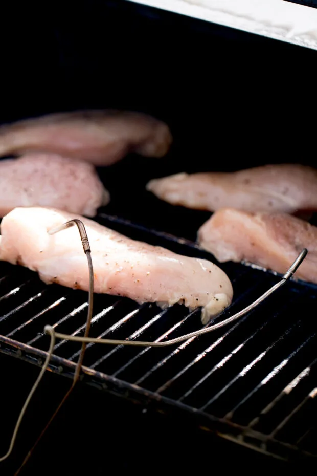 Raw chicken breasts on the grates of a pellet grill with temperature probes inserted into two of them.