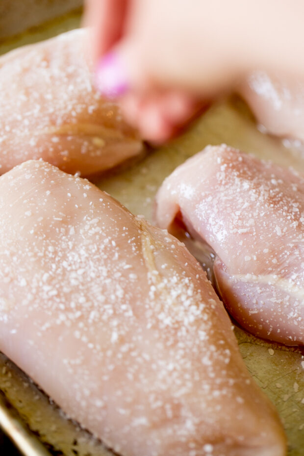 Raw chicken breasts on a sheet pan being sprinkled with kosher salt