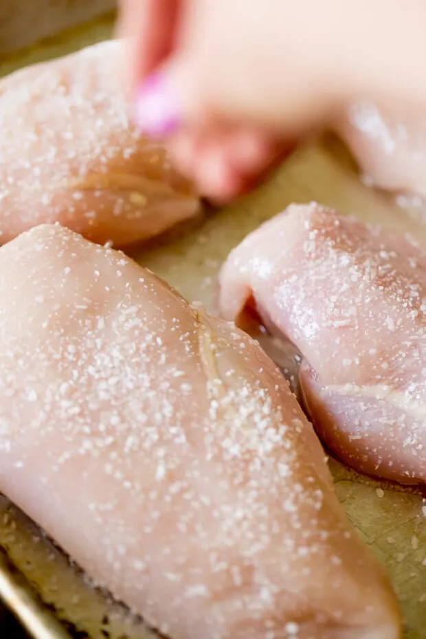 Raw chicken breasts on a sheet pan being sprinkled with kosher salt