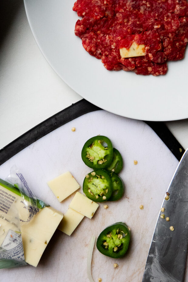 A white plate with an uncooked beef patty filled with pepperjack cheese chunks. Next to it is a white cutting board with sliced jalapeno peppers and pepperjack cheese. 