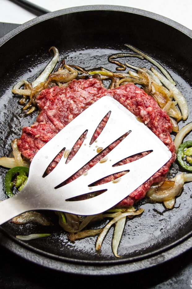 A beef patty is pressed into caramelized onions and jalapenos in a cast iron skillet by a large metal spatula.