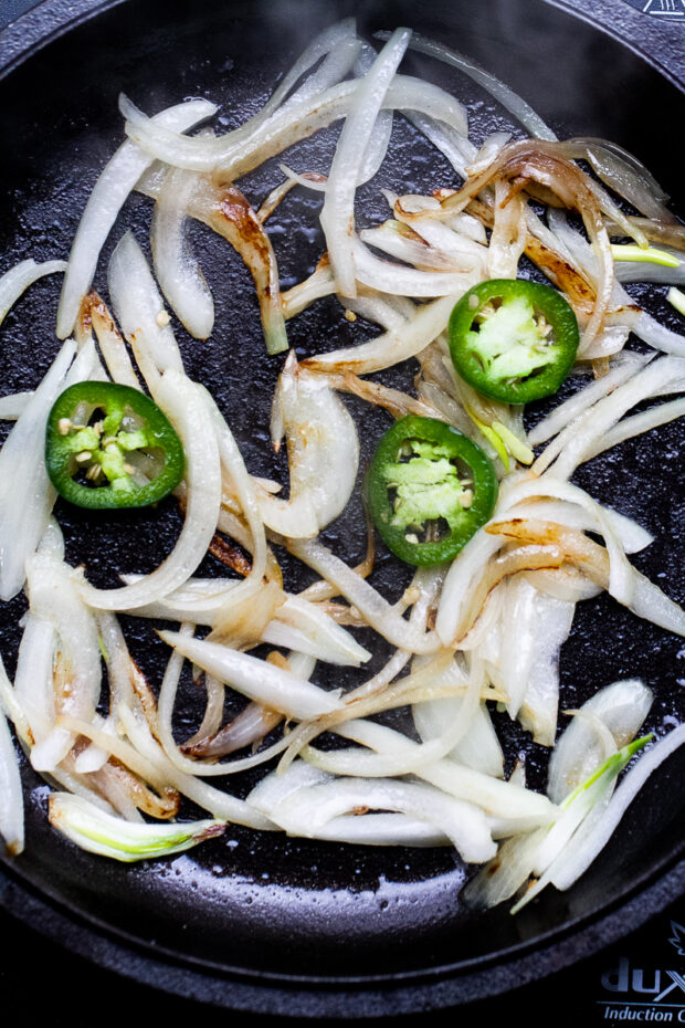 A cast iron skillet with sliced onion and jalapenos starting to brown.