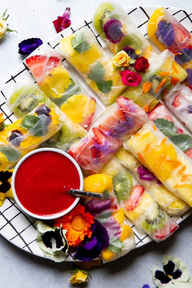 A platter of colorful spring rolls filled with fruit and sweet coconut rice sits next to a small bowl of raspberry sauce. A few edible flowers are scattered over the spring rolls and on the surface next to the platter.
