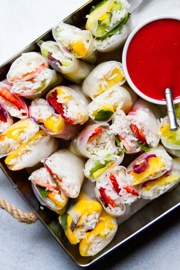A tray with fruity spring rolls cut in half sitting cut-side up next to a small bowl of raspberry sauce.