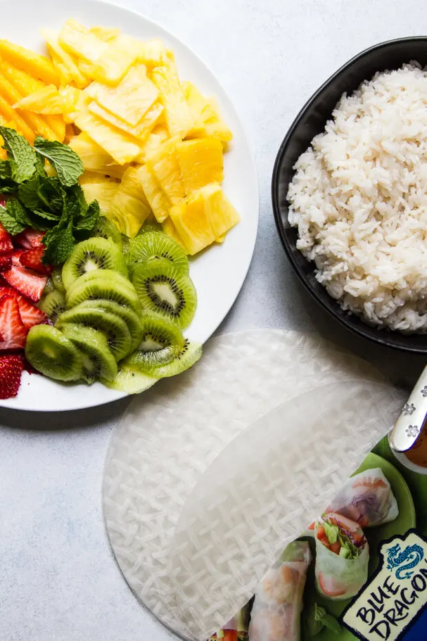 A bowl of sliced fruit sits next to a bowl of sweet coconut rice and an open package of spring roll wrappers.