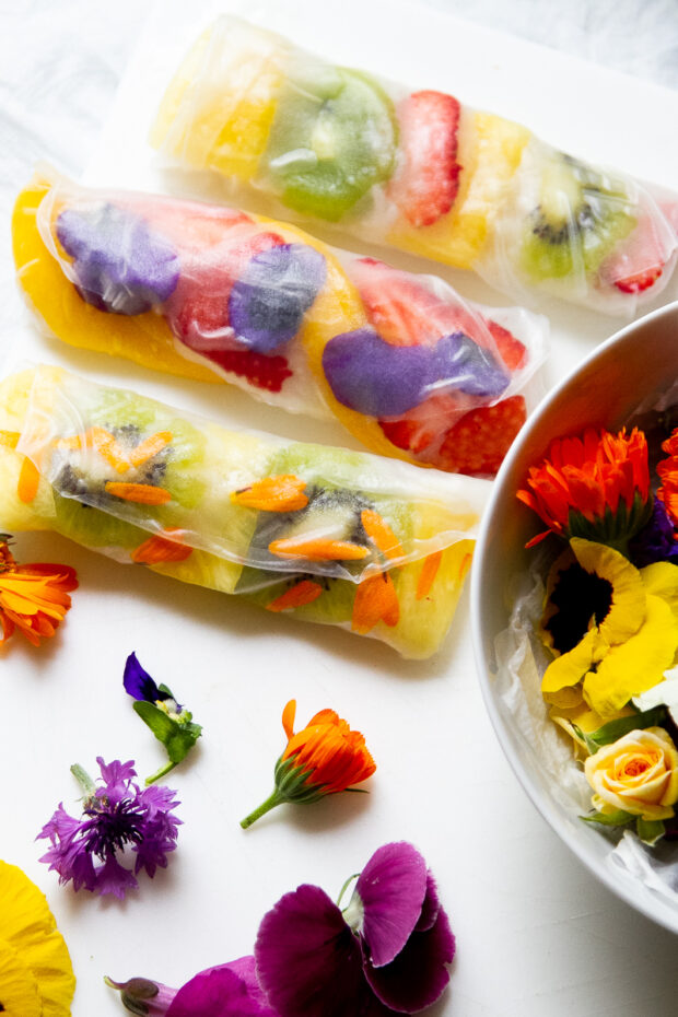 A few fruity spring rolls with edible flowers sitting next to a bowl with more flowers.