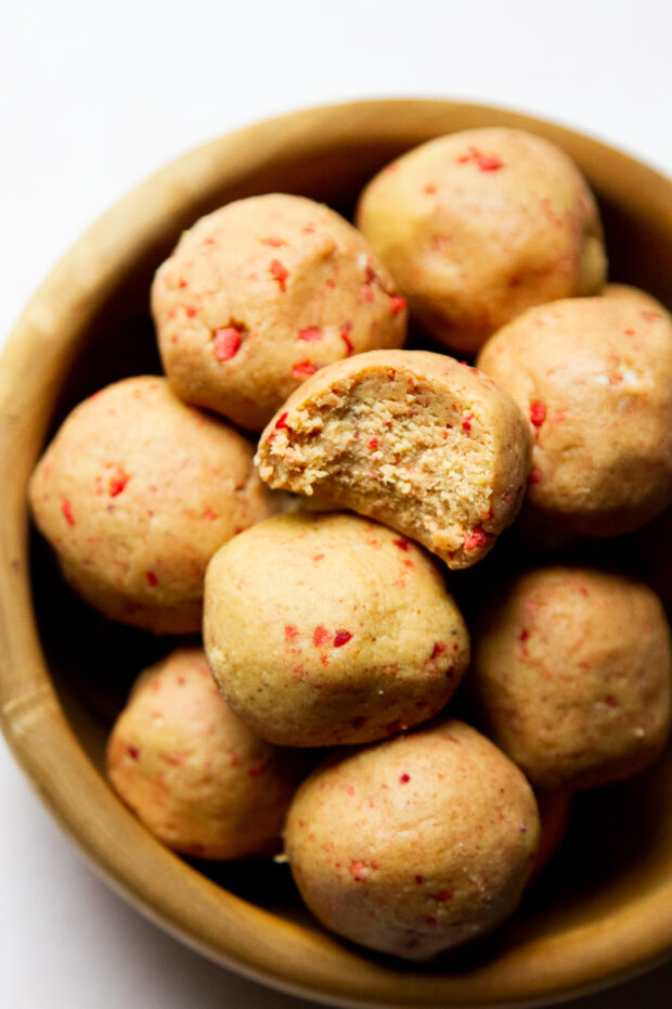 Peanut Butter Protein Bites with bits of freeze-dried strawberries in a wooden bowl. The protein ball on the top has a bite taken out.
