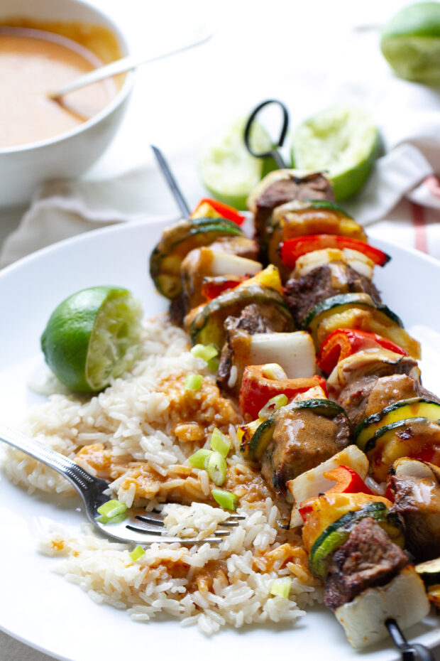 Two Thai steak kebabs on a plate with coconut rice and drizzled with coconut curry sauce.