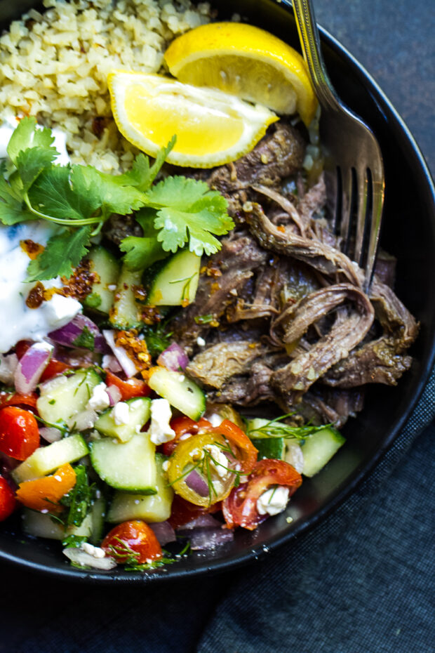 Finished Gyro-inspired shredded beef in a black bowl with roasted cauliflower rice, homemade tzatziki, cucumber tomato salad, some fresh cilantro, and lemon wedges.