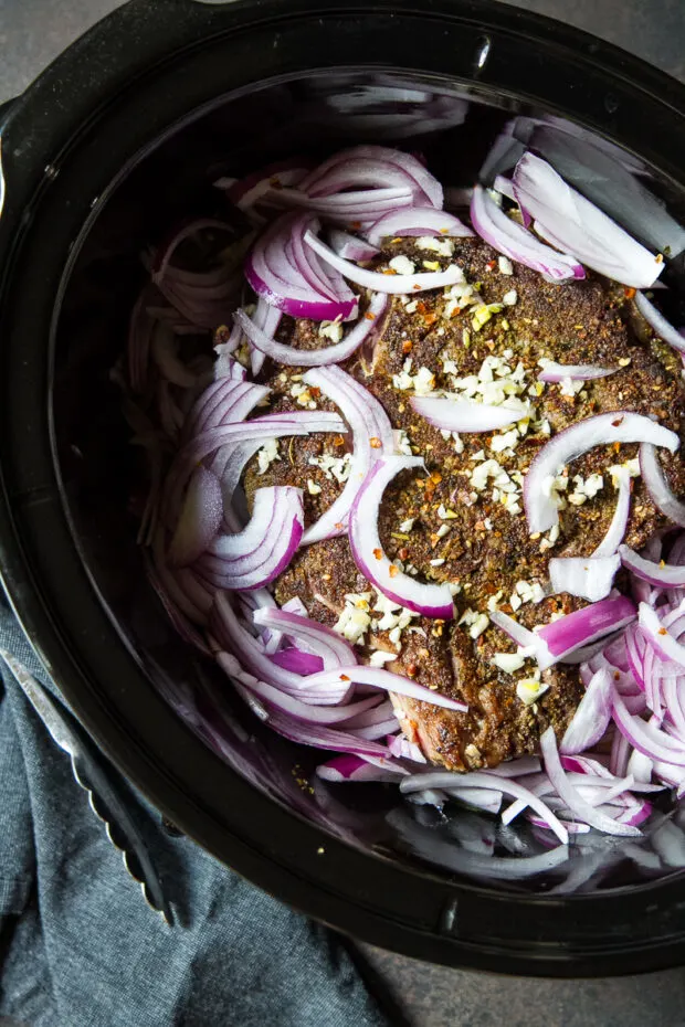 Beef roast in a Crock Pot slow cooker rubbed with Middle Eastern seasoning, seared and topped with fresh garlic and thinly sliced red onion.