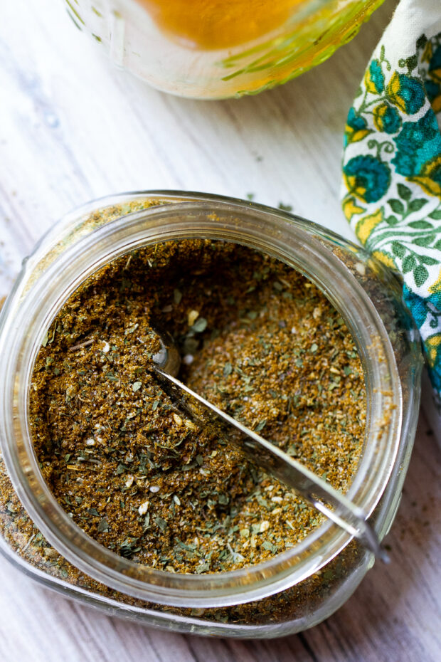 A short, wide-mouth glass jar with Middle Eastern Seasoning blend. A small measuring spoon is stuck inside the blend.