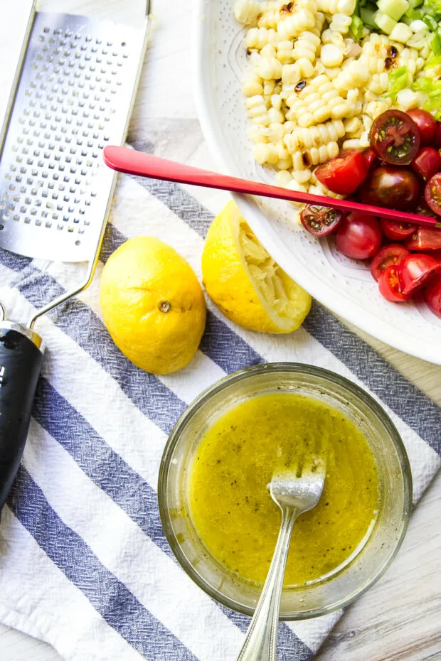 Lemon-garlic vinaigrette in a small glass bowl sitting next to the grilled vegetable salad ready to be dressed. Also pictured is a lemon cut in a half and squeezed next to a microplane grater.