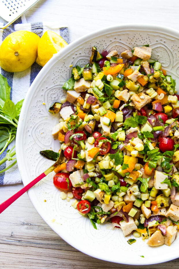 Grilled vegetable and rotisserie chicken salad in a large serving bowl alongside a cut lemon and a sprig of basil.