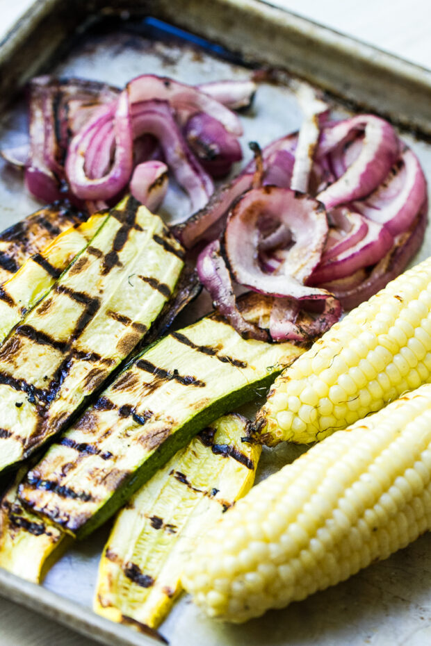 A large rimmed baking sheet with grilled zucchini slices. grilled red onion rings, and a couple of ears of grilled corn.