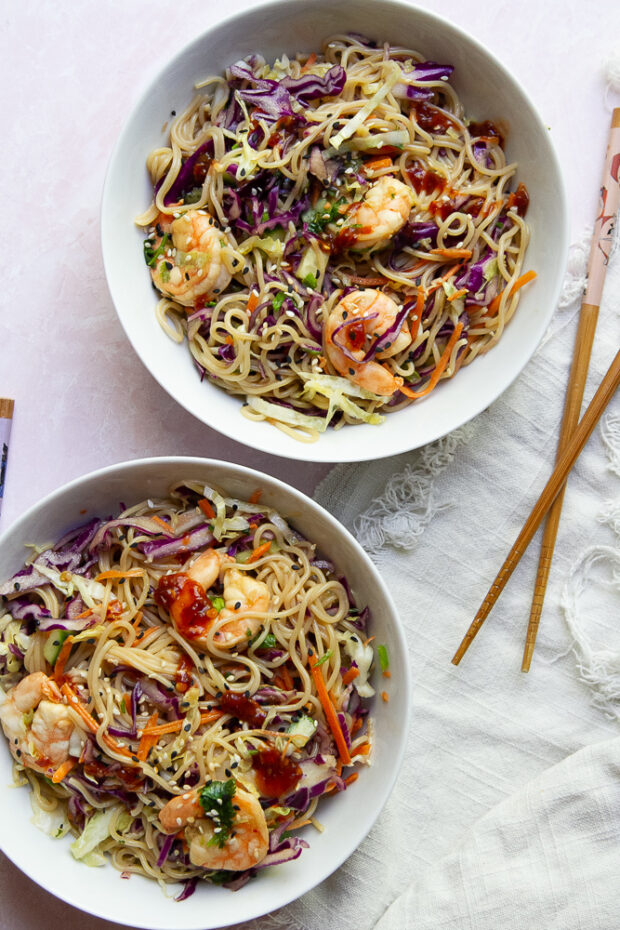 Two servings of the Shrimp & Ramen Noodle Bowls are in shallow white bowls sprinkled with sesame seeds and drizzled with sriracha. Chopsticks sit alongside ready to be used.