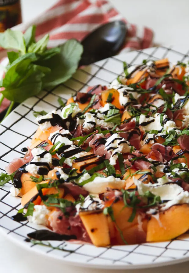 Another shot of the finished cantaloupe, prosciutto, and burrata salad next to a tea towel and a sprig of fresh basil.