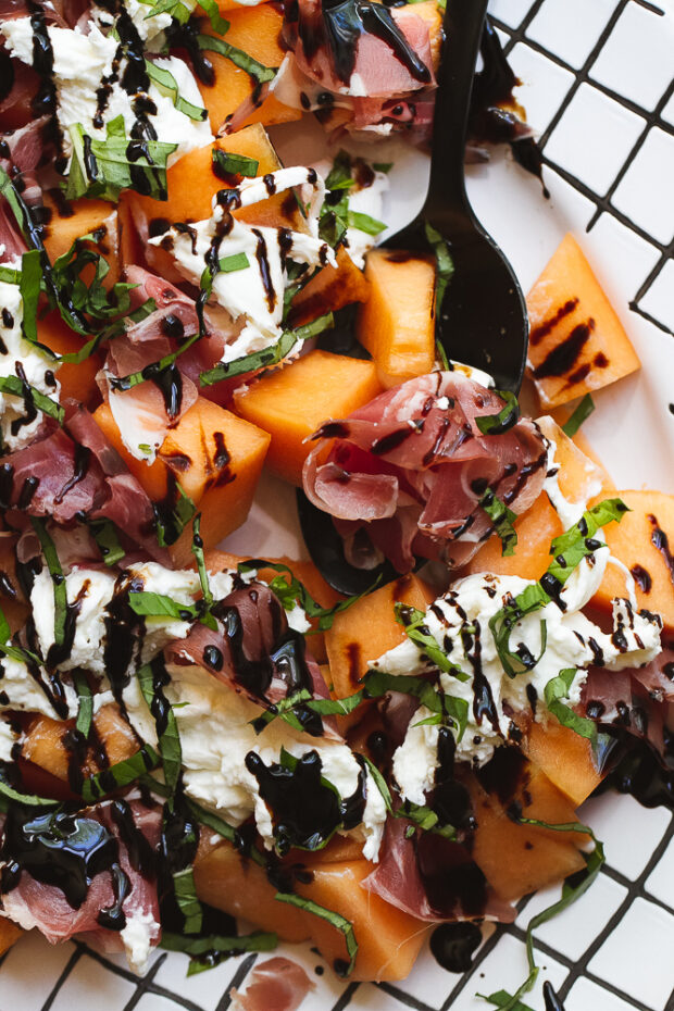 Cantaloupe, prosciutto, & burrata salad drizzled with balsamic glaze and sprinkled with fresh basil. The salad is on a white platter with black cross-hatching around the edges.