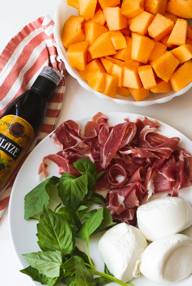 Ingredients for this salad -- cubed cantaloupe in a bowl, balsamic glaze, and a plate with ripped prosciutto, a sprig of fresh basil, and some burrata cheese.