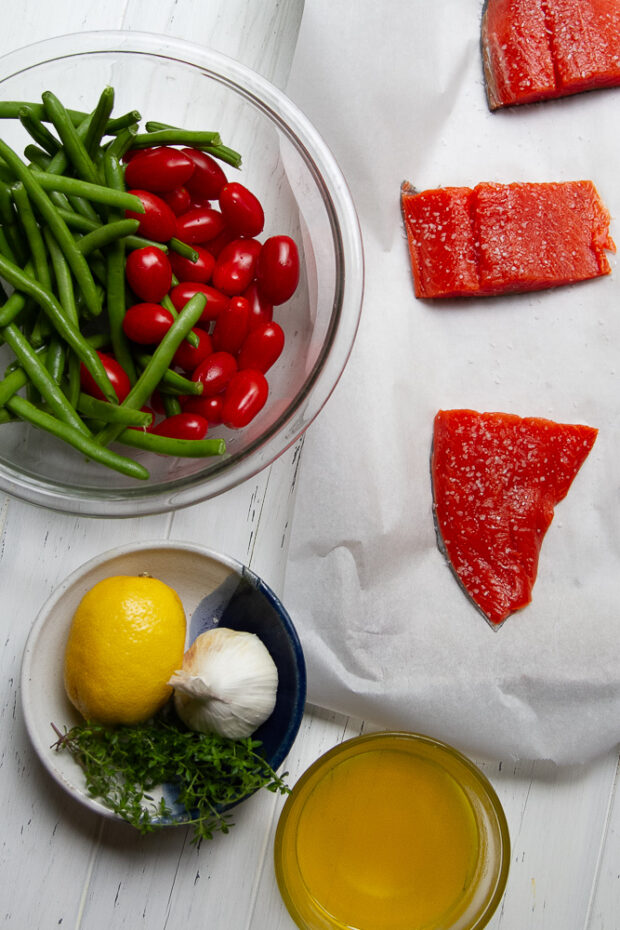 The ingredients for this recipe laid to on a table -- uncooked sockeye salmon fillets, fresh green beans and grape tomatoes, melted butter, fresh garlic, lemon, and thyme sprigs.