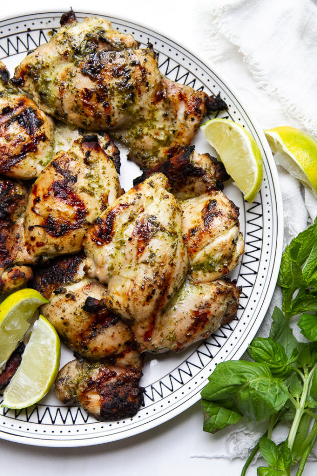 Finished grilled chicken thighs with herby lime marinade on a platter with lime wedges and springs of fresh herbs.