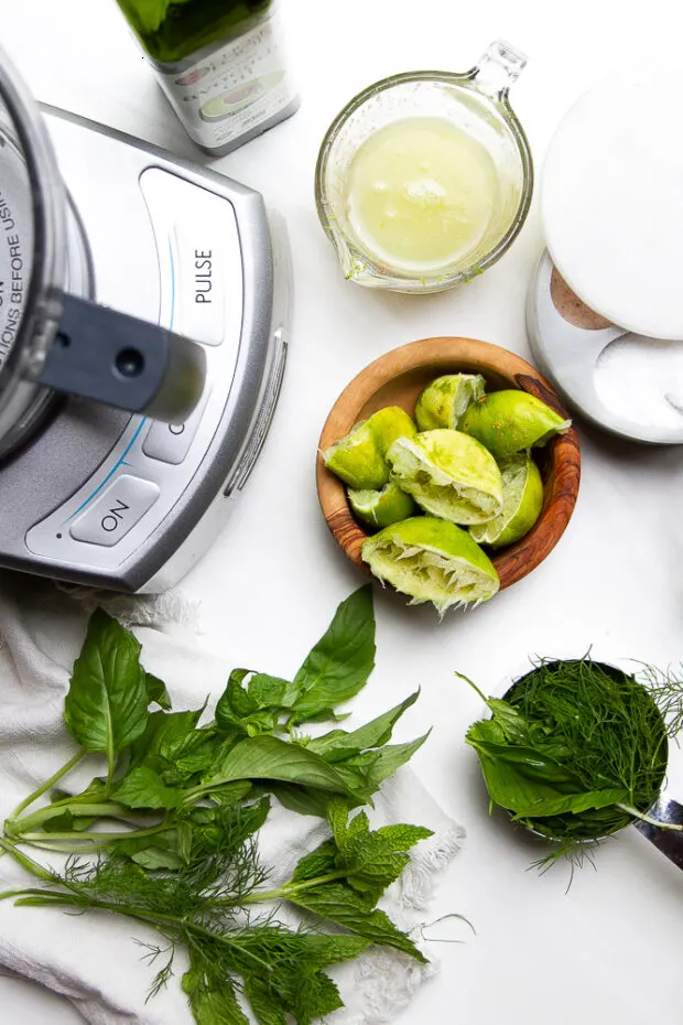 Ingredients for the lime herb marinade ready to put in the food processor. A glass measuring cup full of freshly squeezed lime juice, a bowl of squeezed out limes, a salt cellar, and a measuring cup packed with fresh herbs.