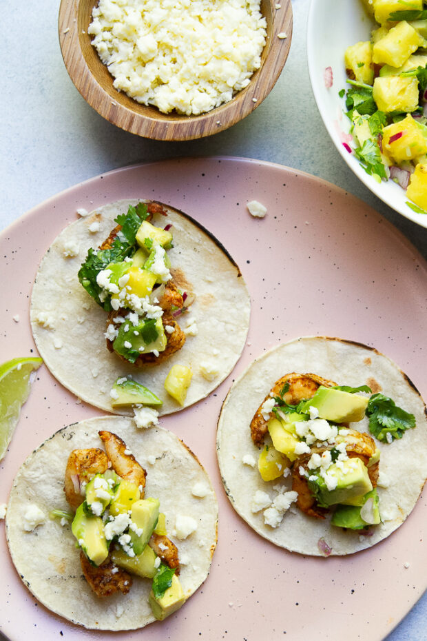 Three grilled shrimp tacos with pineapple avocado salsa and a sprinkle of Mexican cotija cheese are on a pale pink plate next to the garnishes in bowls.