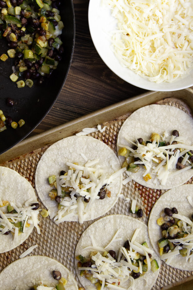 Prepping the black bean tacos. A sheet pan shows street corn tortillas flat with a little filling and some shredded pepper jack cheese. Ready to go in the oven.