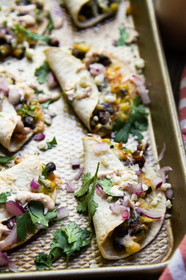 Crispy Black Bean Tacos on a sheet pan fresh from the oven topped with red onion, fresh cilantro, and a drizzle of creamy chipotle sauce.