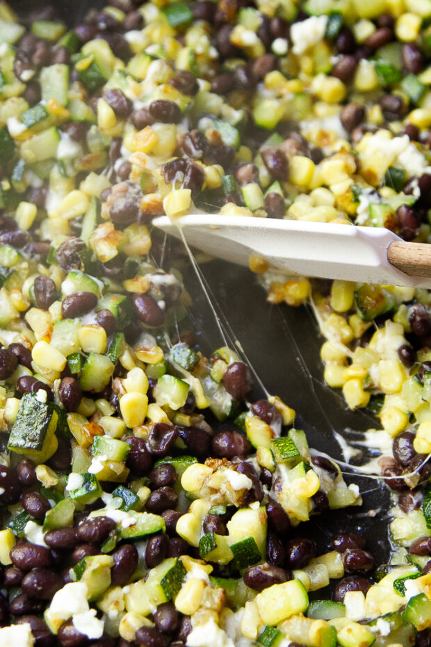 View of the black beans, corn, and diced zucchini sautéed in a pan with garlic butter. Melted cotija cheese connects the tender bits with strings.