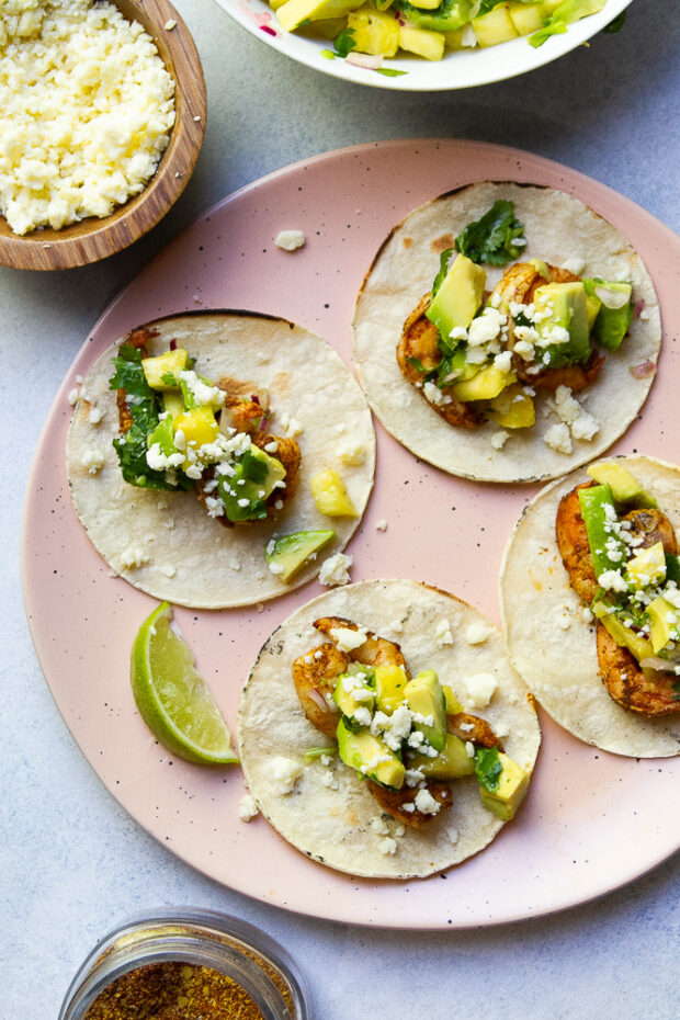 The same pink plate as above with 4 assembled grilled shrimp tacos and pineapple avocado salsa and a lime wedge along the side.