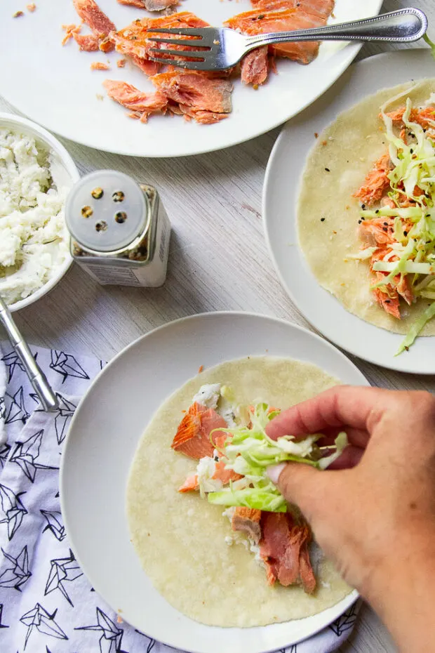 Assembling the smoked salmon taco by smearing some herb cheese spread in the center of the taco, adding some salmon, and some shredded cabbage.