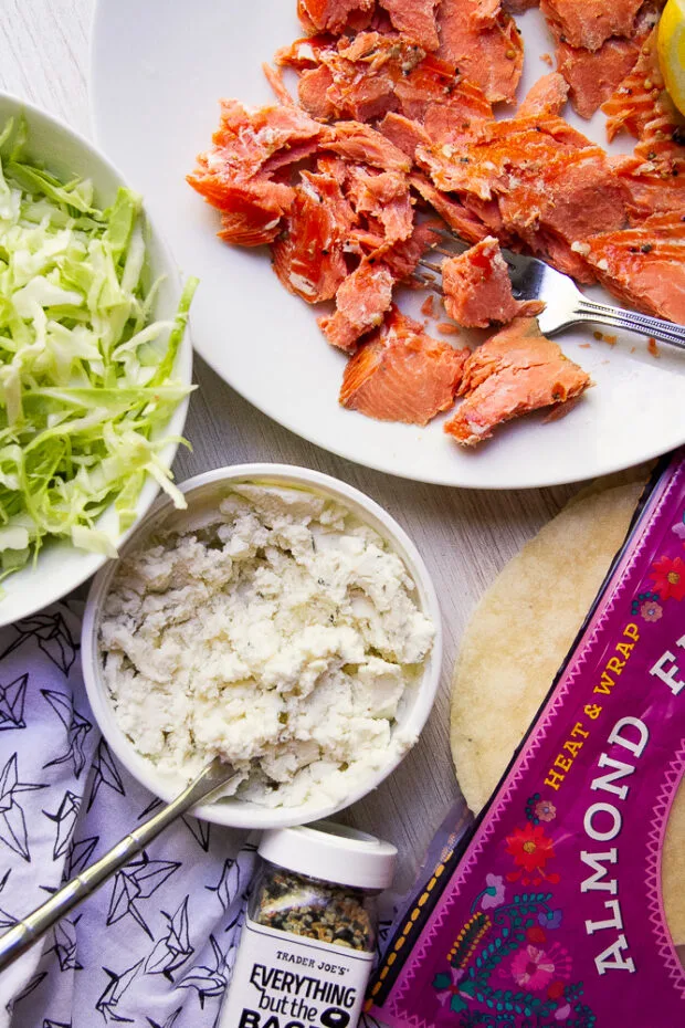 Ingredients sitting out for smoked salmon tacos: A plate of broken up hot smoked salmon, small almond flour tortillas, Everything Bagel Seasoning, herb cheese spread, and shredded cabbage.