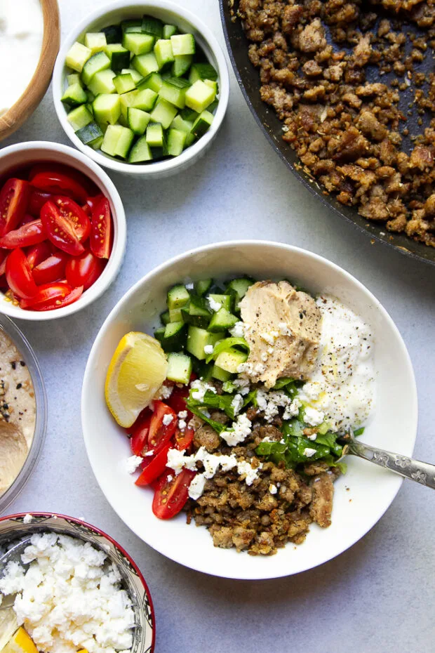 Components for the ground turkey bowls laid out for assembly. A finished bowl is shown surrounded by the skillet with the ground turkey and small bowls containing diced cucumbers and halved cherry tomatoes.