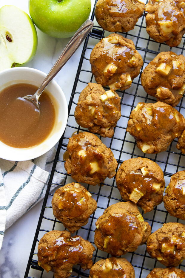 Shot of glazed gluten free apple cookies on a cooling rack next to a small bowl of maple glaze and a cut green apple.