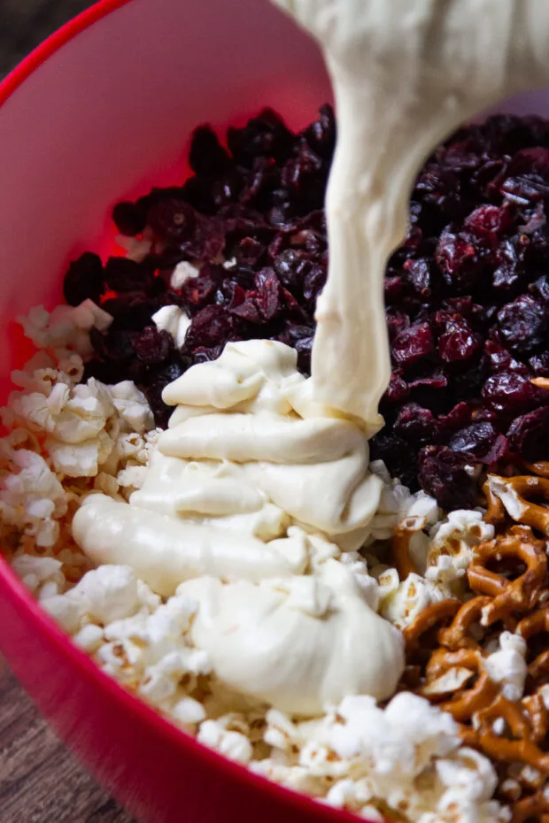 Melted white chocolate poured over a very large bowl with air popped popcorn, dried cranberries, and gluten-free pretzels.